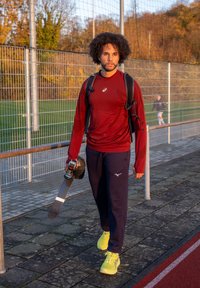 Atletisk outfit med en mørkerød langærmet skjorte, marineblå bukser og lysegule sneakers. Bærer et skateboard og har en rygsæk på.