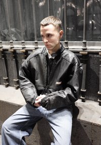 Giacca bomber nera in pelle con chiusura a zip, collo a costine e vestibilità ampia, abbinata a jeans larghi azzurri, seduta contro delle sbarre di metallo.