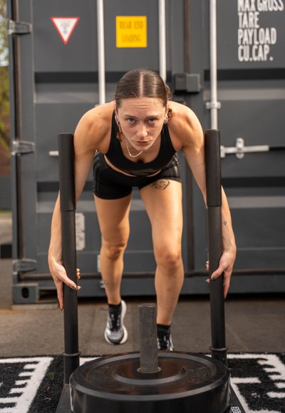 Trineo negro pesado con asas verticales, empujado por una persona con una camiseta sin mangas negra y pantalones cortos, sobre una superficie exterior texturizada.