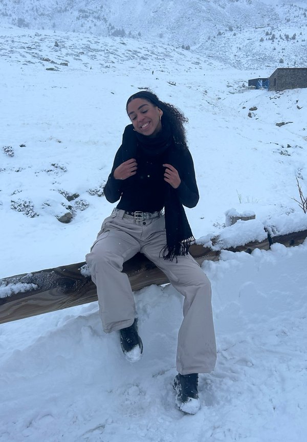 Tenue décontractée comprenant un haut noir à manches longues, une écharpe noire, un pantalon de neige crème et des bottes noires. Fond de paysage enneigé.
