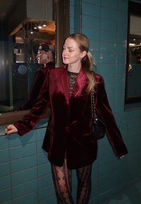 Bordeauxrode fluwelen blazer met oplopende revers, gecombineerd met een doorschijnende zwarte kanten top en zwarte panty's met patroon. Zwarte handtas met ketting hengsel.