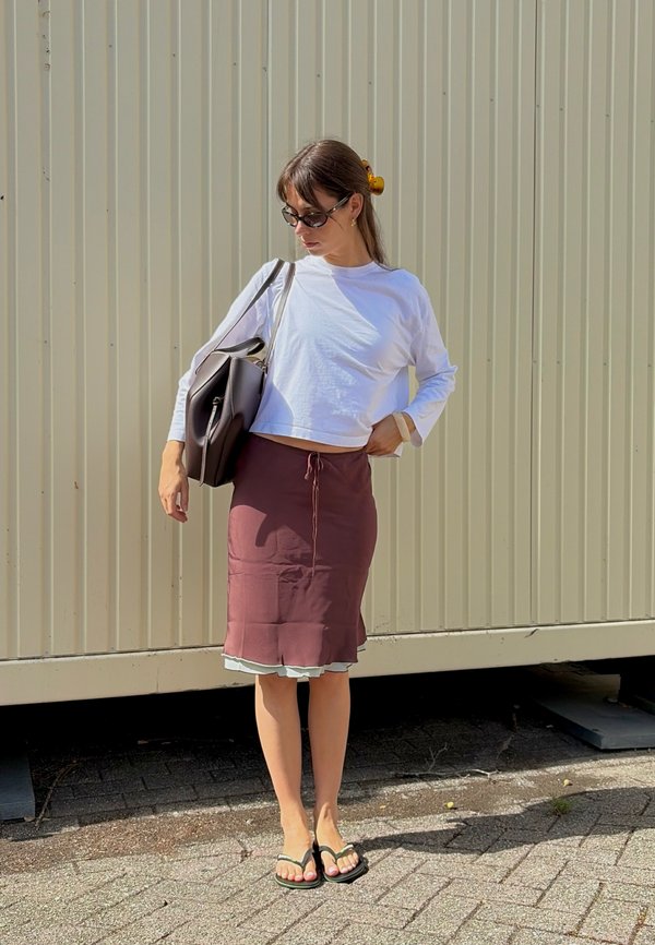 Femme portant un haut blanc à manches longues, une jupe bordeaux, des sandales noires, des lunettes de soleil et portant un grand sac noir, debout contre un mur ondulé.