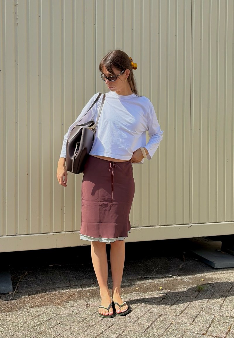 Femme portant un haut blanc à manches longues, une jupe bordeaux, des sandales noires, des lunettes de soleil et portant un grand sac noir, debout contre un mur ondulé.