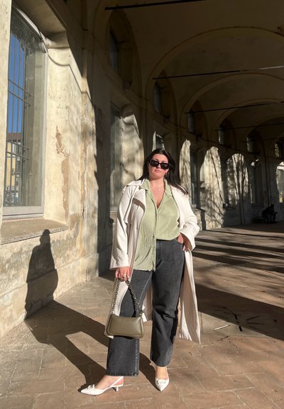 Camisa de rayas verde claro, abrigo trench color crema, jeans de pierna ancha negros, tacones blancos de punta y un bolso verde oliva sobre un fondo texturizado.