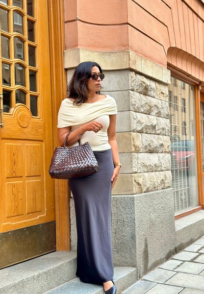 Bolso trenzado marrón, blusa crema sin hombros y falda larga gris. Tejido suave, zapatos negros de punta redonda, de pie cerca de una puerta de madera y una pared de piedra.