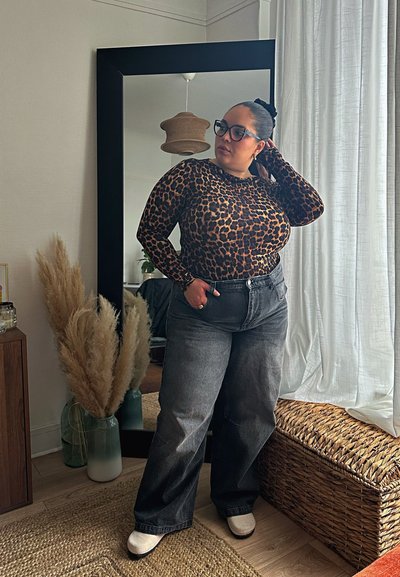 Top de manga larga con estampado de leopardo combinado con jeans de mezclilla gris de cintura alta. Zapatillas blancas, cabello trenzado y un espejo que refleja un ambiente hogareño.