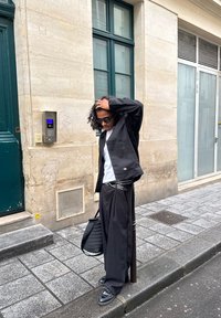 Mężczyzna o kręconych włosach i w okularach przeciwsłonecznych, ubrany w czarną kurtkę, szerokie spodnie i białą koszulę, stoi na chodniku w mieście, trzymając czarną torbę.