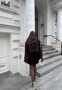 Ruda netikro kailio striukė, permatomos raštuotos pėdkelnės, balti aukštakulniai ir maža metalinė rankinė; dėvima lauke stovinčio žmogaus ant akmeninių laiptų.