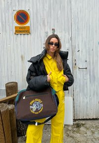 Μαύρο μπουφάν, κίτρινη φόρμα, και καφέ τσάντα με μπλε λεπτομέρεια και κεντημένο σχέδιο. Το φόντο περιλαμβάνει ξύλινες κολόνες και μια λευκή πόρτα.