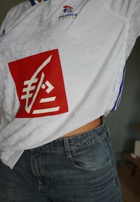 Baltas sportinis marškinėliai su gėlių raštu, raudonas kvadratinis logotipas su baltais grafiniais elementais ir mėlynos juostelės palei pečius. Dėvima su nusidėvėjusiais džinsais.