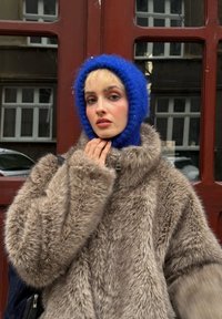 Manteau en fausse fourrure marron clair avec un col montant ; chapeau bleu duveteux ; modèle à la peau pâle et aux cheveux courts et clairs ; arrière-plan urbain.