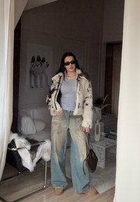 Jakna iz umetnega krzna s pikčastim vzorcem, siva majica, svetlo modre široke kavbojke in rjava torba. Oseba stoji v sodobnem notranjem prostoru.