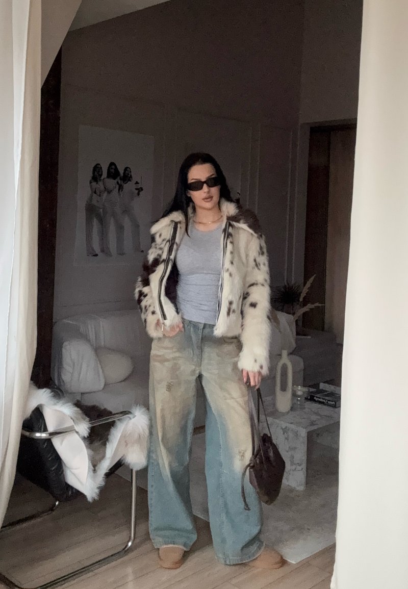Giacca in pelliccia sintetica con motivo a macchie, t-shirt grigia, jeans a gamba larga in denim chiaro e borsa marrone. La persona è in piedi in uno spazio interno moderno.