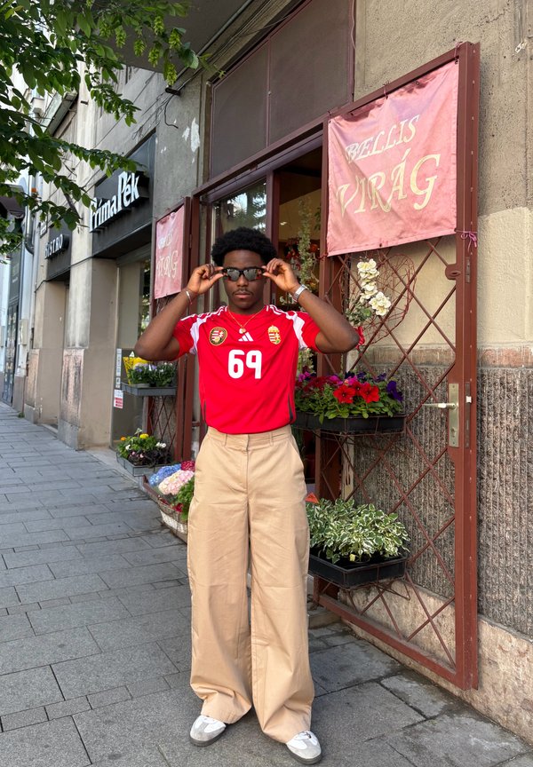 Rotes Fußballtrikot mit "69" Grafik, beigen weitem Hosenanzug und schwarzen Sonnenbrillen. Blumenladen-Hintergrund mit verschiedenen Pflanzen und Blüten.