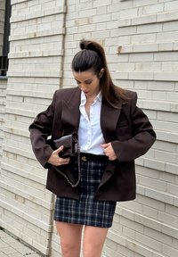 Femme à queue de cheval ajustant un blazer oversize marron foncé sur une chemise blanche et une jupe à carreaux, tenant un sac à main sombre contre un mur en briques claires.