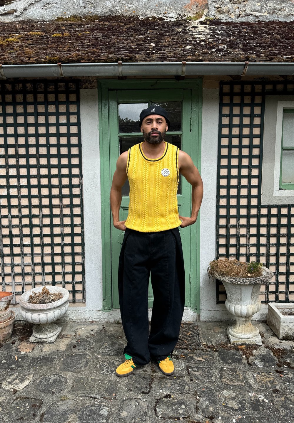 Suéter amarillo de punto sin mangas con cuello redondo, combinado con pantalones negros holgados y zapatillas amarillas. El fondo presenta una puerta verde.