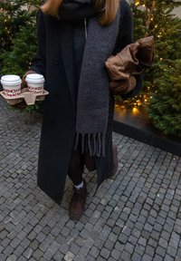 Personne portant un manteau sombre, une écharpe et des chaussures marron, tenant deux tasses de café et des gants marron, debout sur des pavés près de sapins de Noël illuminés.