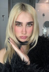 Femme blonde aux yeux verts, lèvres brillantes et ongles noirs décorés, posant avec la main près du visage, vêtue d'un vêtement noir duveteux dans une pièce carrelée.