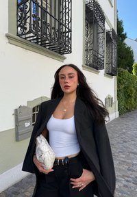 Zwarte blazer, wit tanktop en zwarte broek. Houdt een creme kleurige, gevlochten clutch vast. Omgeven door een kiezelstenen pad en een witte muur met ventilatieopeningen.