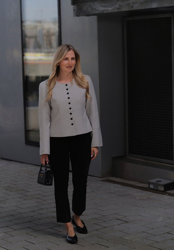 Žena s dugom plavom kosom koja nosi sivu jaknu s kopčama, crne hlače i crne cipele, nosi malu crnu torbicu na otvorenom.