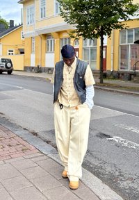 Ung person som bär en svart läderväst över en rutig kortärmad skjorta, creamfärgade byxor och bruna stövlar, stående på en trottoar.