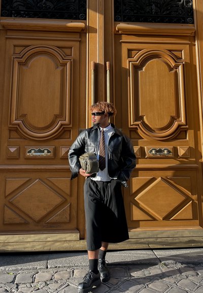 Chaqueta de cuero negra corta, camisa blanca con corbata, falda negra hasta la rodilla y zapatos negros. Sosteniendo un bolso verde. Puertas dobles de madera al fondo.