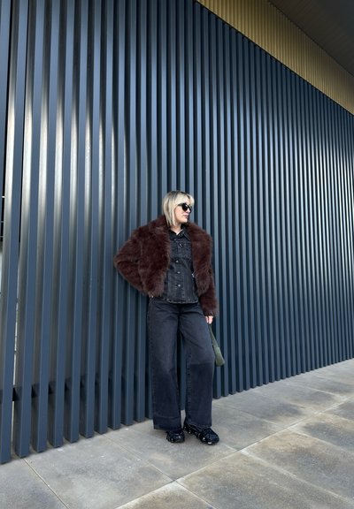 Modelo con una chaqueta de mezclilla oscura y un cuello de piel sintética marrón, pantalones anchos negros y calzado brillante negro, frente a una pared de rayas verticales.