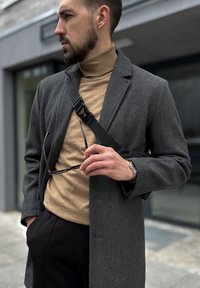 Abrigo de lana gris sobre un suéter de cuello alto beige, sosteniendo gafas de sol. Correa negra cruzada sobre el hombro, reloj visible en la muñeca. Escenario urbano moderno.