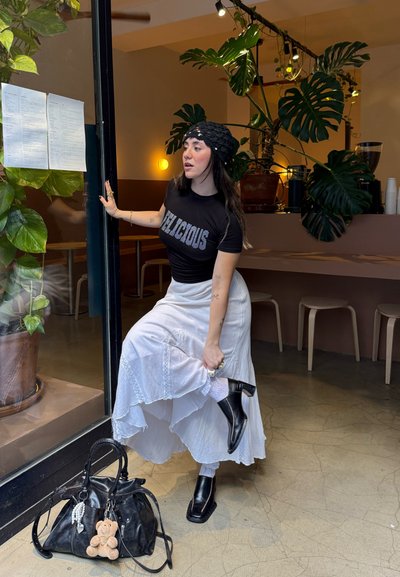 Camiseta negra con la palabra "DELICIOUS" en strass, falda blanca fluida, botas negras hasta el tobillo y un bolso negro texturizado. Plantas verdes de fondo.