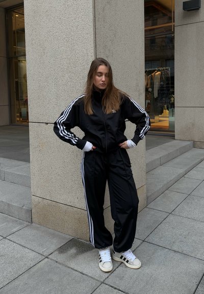 Chándal negro con rayas blancas, chaqueta con cremallera y pantalones a juego, combinados con zapatillas blancas con detalles en negro, de pie al aire libre.