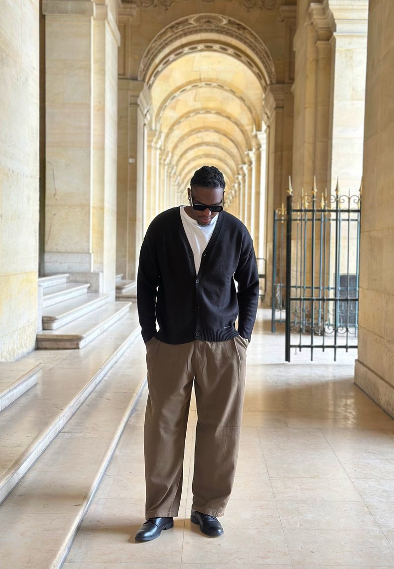 Homme portant des lunettes de soleil, un cardigan noir, une chemise blanche et un pantalon marron, debout les mains dans les poches sous un corridor voûté en pierre.