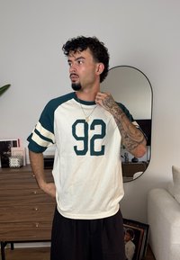 T-shirt bianco e teal con grafica 92. Presenta maniche raglan, una vestibilità rilassata e un design a righe contrastanti sulle maniche.