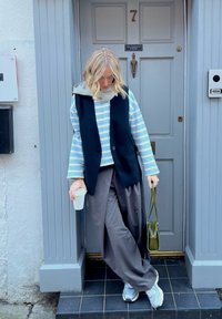 Model v pruhovaném modro-bílém topu, černé bezrukávové vestě, šedých širokých kalhotách, držící šálek kávy a zelenou kabelku. Na nohou tenisky.