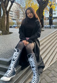 Schwarzer langer Mantel über einem schwarzen Rollkragenpullover, kombiniert mit kniehohen schwarzen Stoffstiefeln mit weißen Schnürsenkeln, sitzend auf Steinstufen mit Kies.