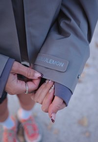 Rokas ar dekorētajiem nagiem pielāgo pelēkā Lululemon jakas apkakli, kas uzvilkta virs purpursarkanas piedurknes, ar izplūdušām kurpēm fona.