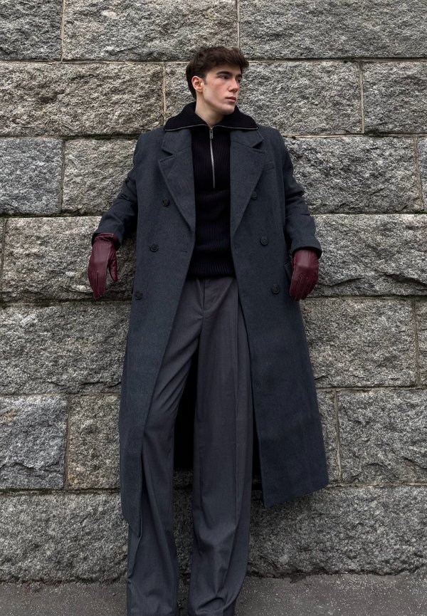Long manteau gris foncé à double boutonnage avec de larges revers, associé à un pull noir côtelé et à un pantalon gris plissé ; gants en cuir bordeaux.