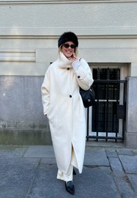 Femme portant un long manteau blanc, un chapeau en fourrure noire, des lunettes de soleil, un sac à main noir et des chaussures noires pointues, debout sur un trottoir en pierre près d'un bâtiment.