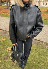 Črni usnjeni suknjič z teksturirano površino, z zadrgo in gumbi. Kombiniran s črnimi širokimi jeans hlačami in usnjeno torbo tote.