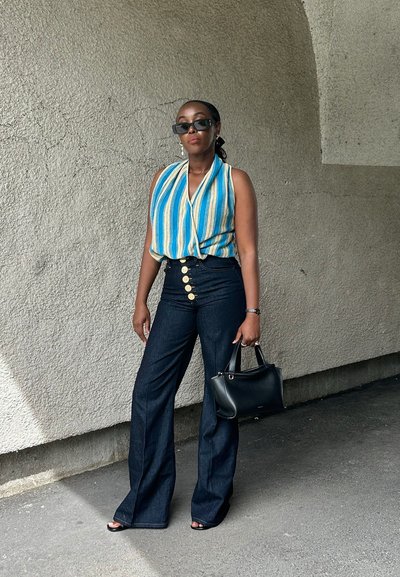 Mujer con top sin mangas a rayas azul y beige, vaqueros oscuros de talle alto con grandes botones, gafas de sol negras y sosteniendo un bolso negro.