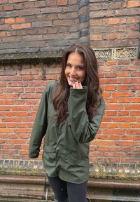 Giacca impermeabile verde con chiusura a bottoni, maniche lunghe e tasche frontali, indossata sopra pantaloni neri, su sfondo di muro di mattoni.