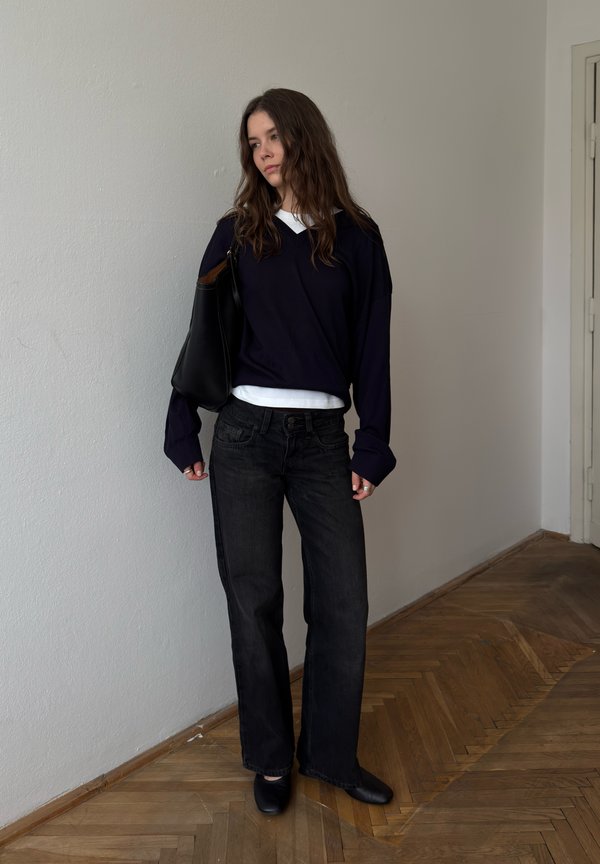 Junge Frau mit langen Haaren, die einen dunklen Pullover, schwarze Jeans und schwarze Schuhe trägt, steht drinnen vor einer weißen Wand mit einer schwarzen Umhängetasche.