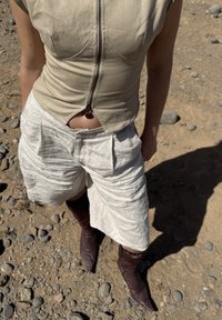 Person iført beige ærmeløs top med lynlås foran, hvide bukser med vide ben og brune spidse støvler, stående på klippefyldt jord.
