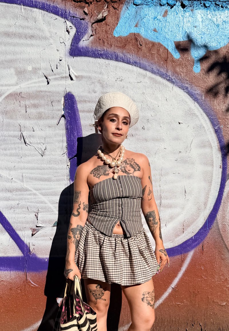 Persona con tatuajes que lleva un top tipo tubo a rayas, falda de cuadros, collar de perlas, boina blanca, sosteniendo un bolso a rayas, de pie frente a una pared con grafitis.