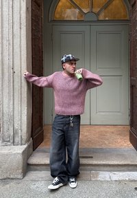 Paks roosa soonik, tume laiad püksid, mustad ja valged sneakerid, hoides roheline jooki, seismine helesinise ukse juures.