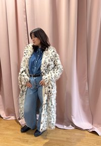 Femme portant un long manteau en fourrure tachetée sur une chemise en denim et un jean large, debout sur un sol en bois avec des rideaux roses en arrière-plan.