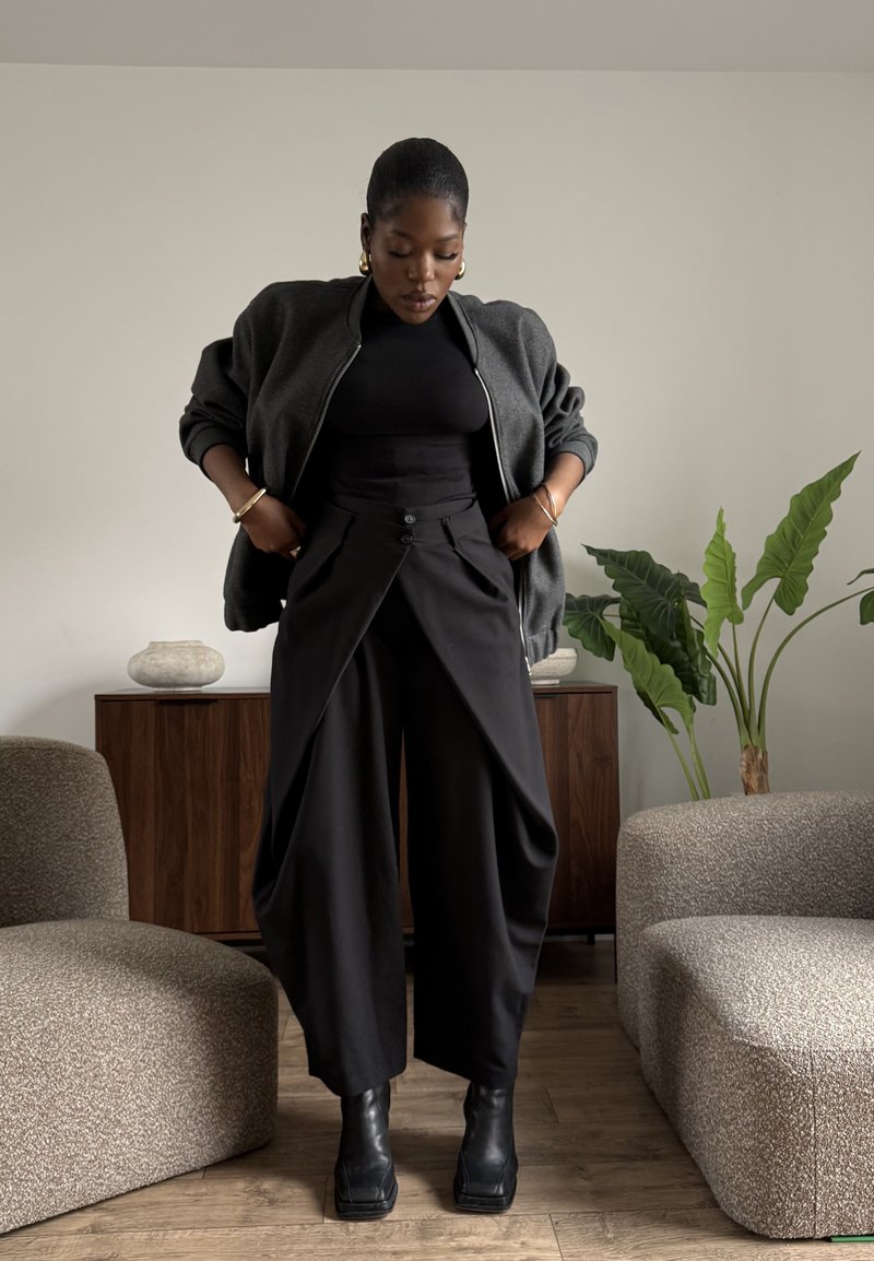 Femme ajustant un pantalon noir à jambes larges, portant un haut noir, une veste gris foncé et des bottes noires dans un salon moderne avec des plantes et des meubles.