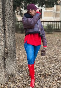 Jachetă maro din piele scurtă cu margine din blană neagră, pulover roșu tricotat, blugi albaștri și cizme de cauciuc roșii, ținând o geantă mică maro, în aer liber.