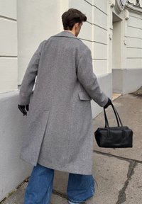 Casaco de lã cinza espinha de peixe com lapelas largas, bolsos laterais e abertura nas costas, combinado com jeans de perna larga e uma pequena bolsa de couro preta.