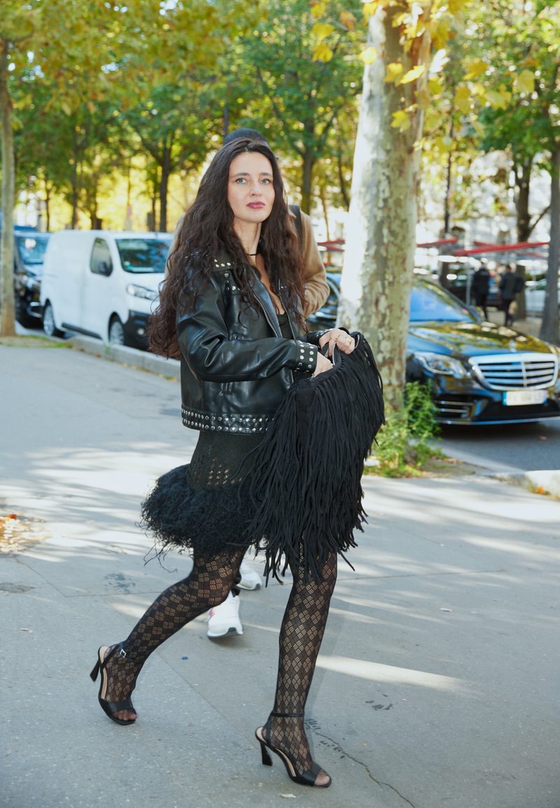 Femme aux longs cheveux noirs portant une veste en cuir cloutée noire, une jupe texturée noire, des collants à motifs et des talons marchant sur le trottoir de la ville.