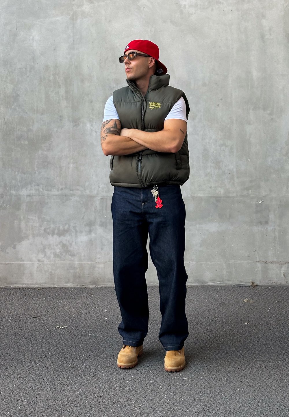 Gilet imbottito verde oliva sopra una camicia bianca, jeans blu scuro, stivali da lavoro color cammello, berretto rosso e occhiali da sole. Accessorio portachiavi visibile.